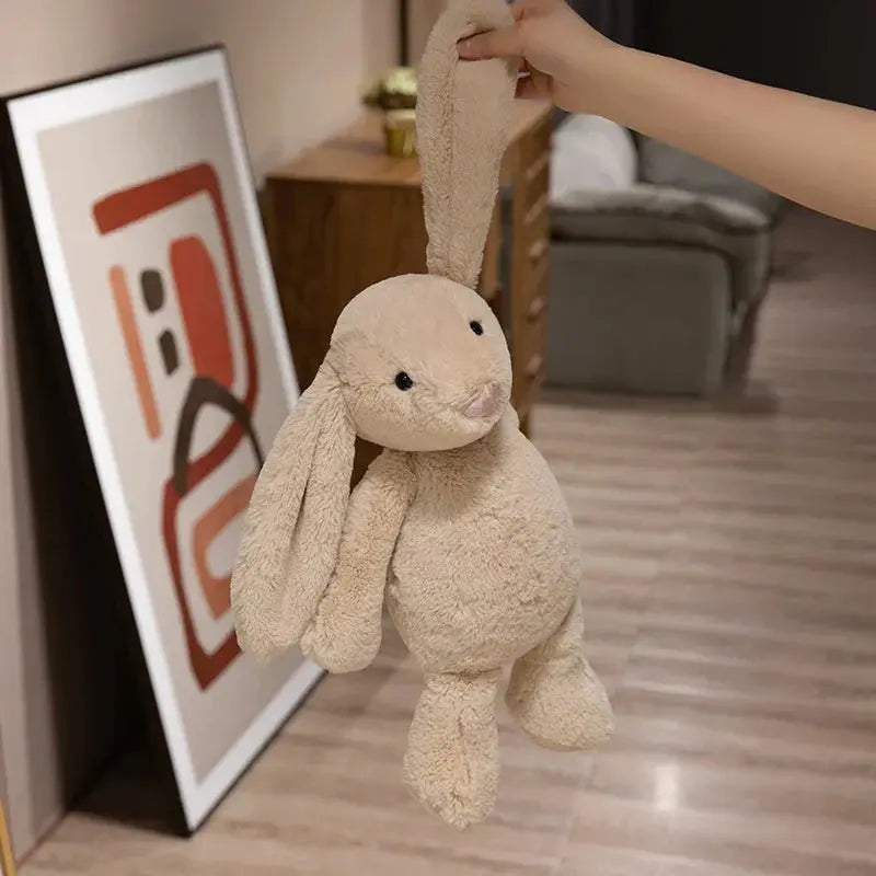 Beige plush bunny held by a hand in a room with a framed picture on the wall.