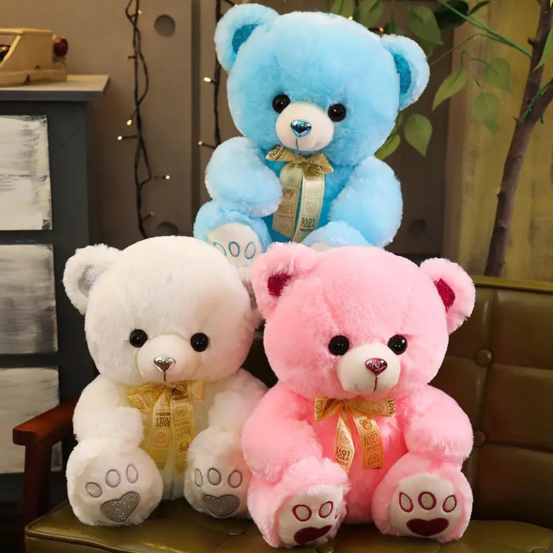 Three Huggable Heart Teddy Bear Plush toys in blue, white, and pink with heart-shaped paw prints and golden bows, displayed on a chair and floor.