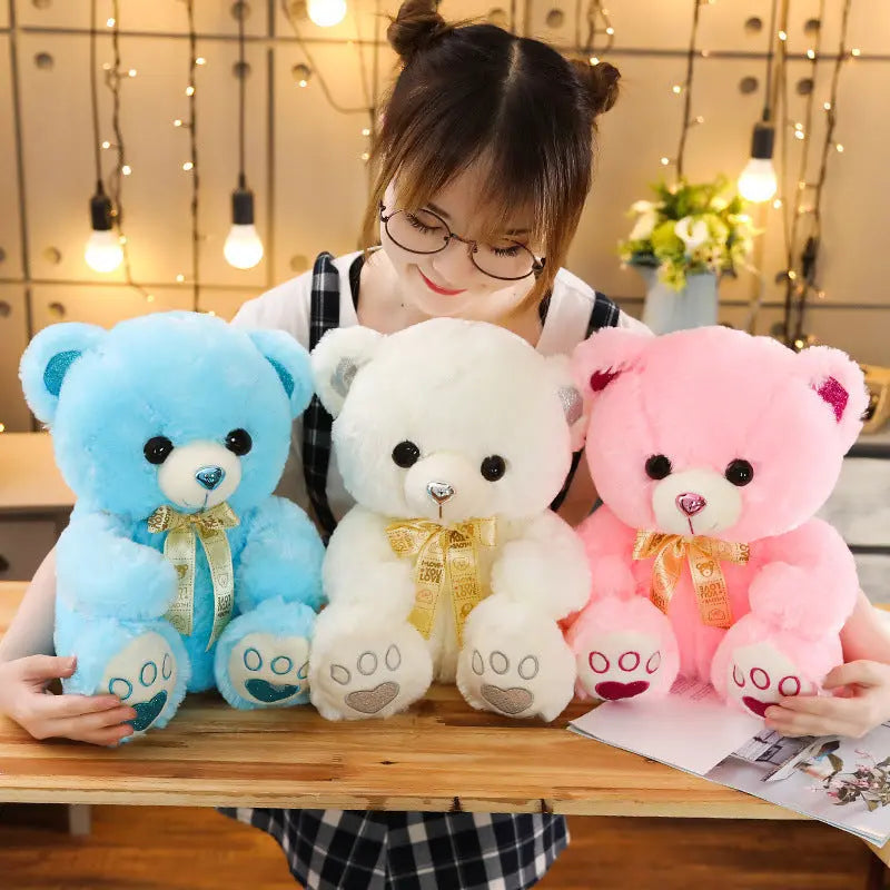 Young woman holding blue, white, and pink Huggable Heart Teddy Bear Plush toys with heart-shaped embellishments on a wooden table.