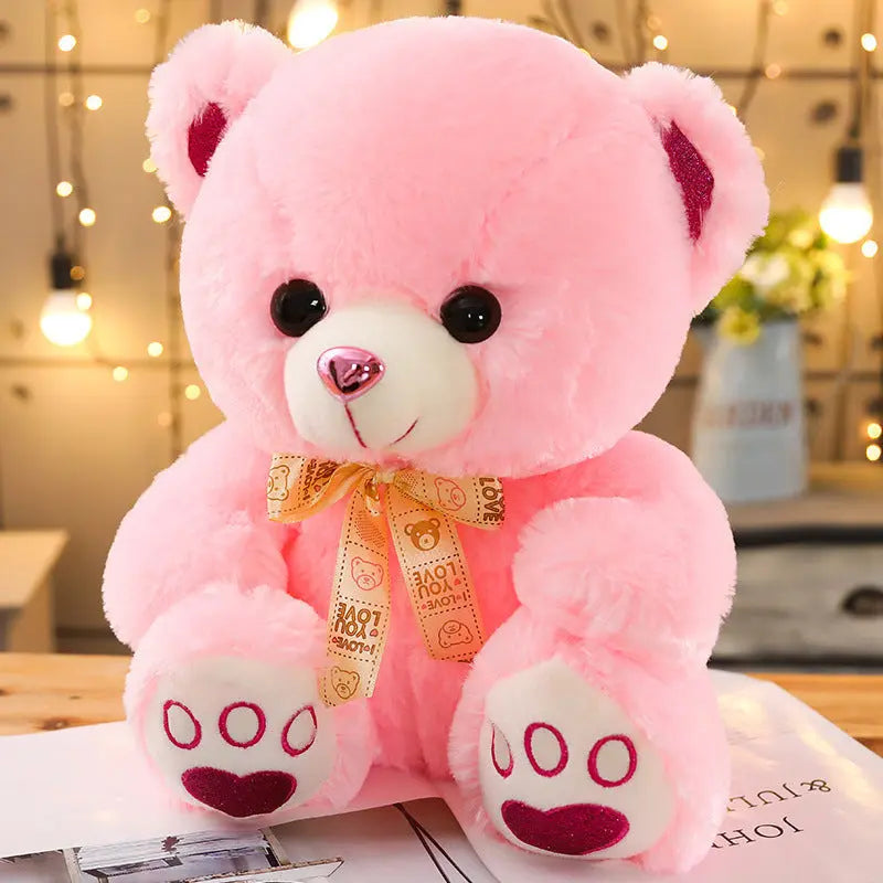 Pink Huggable Heart Teddy Bear Plush with heart-shaped nose and bow ribbon sitting on table with lights background