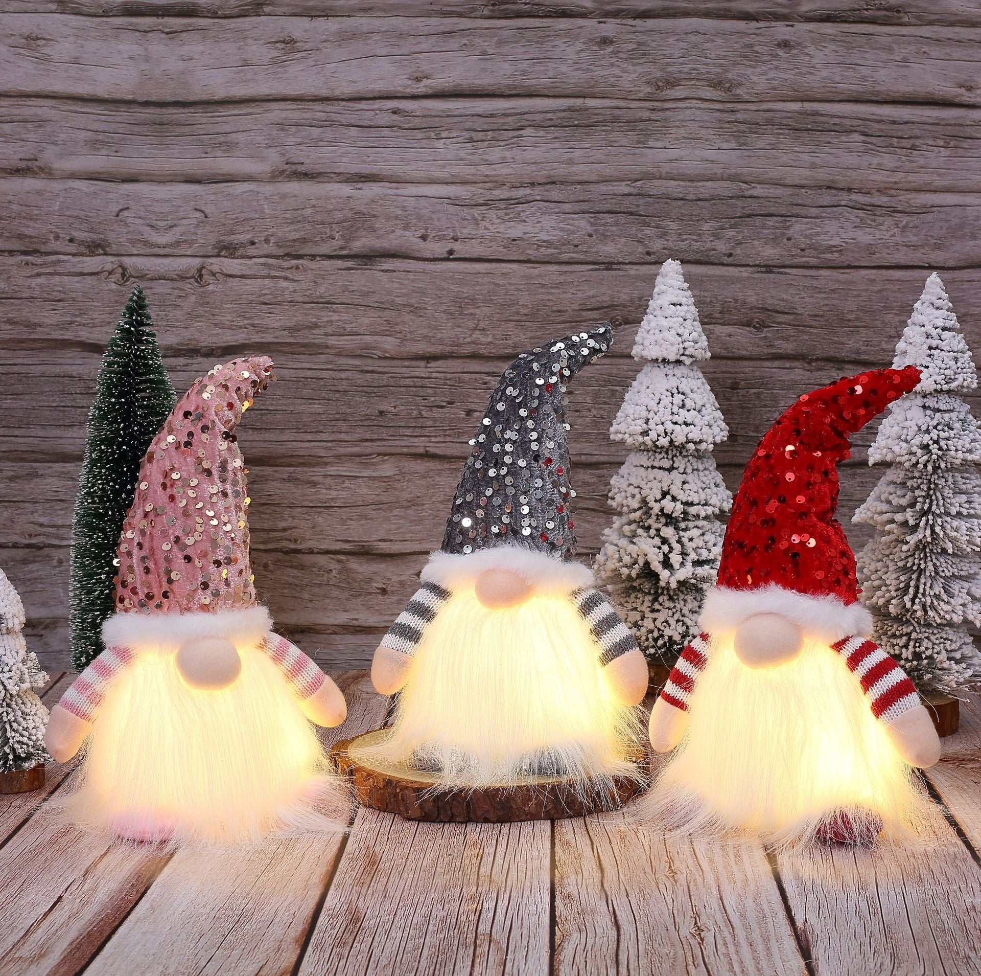 Lighted Christmas gnome decorations with sequined hats in pink, grey, and red on wooden surface with snowy trees background
