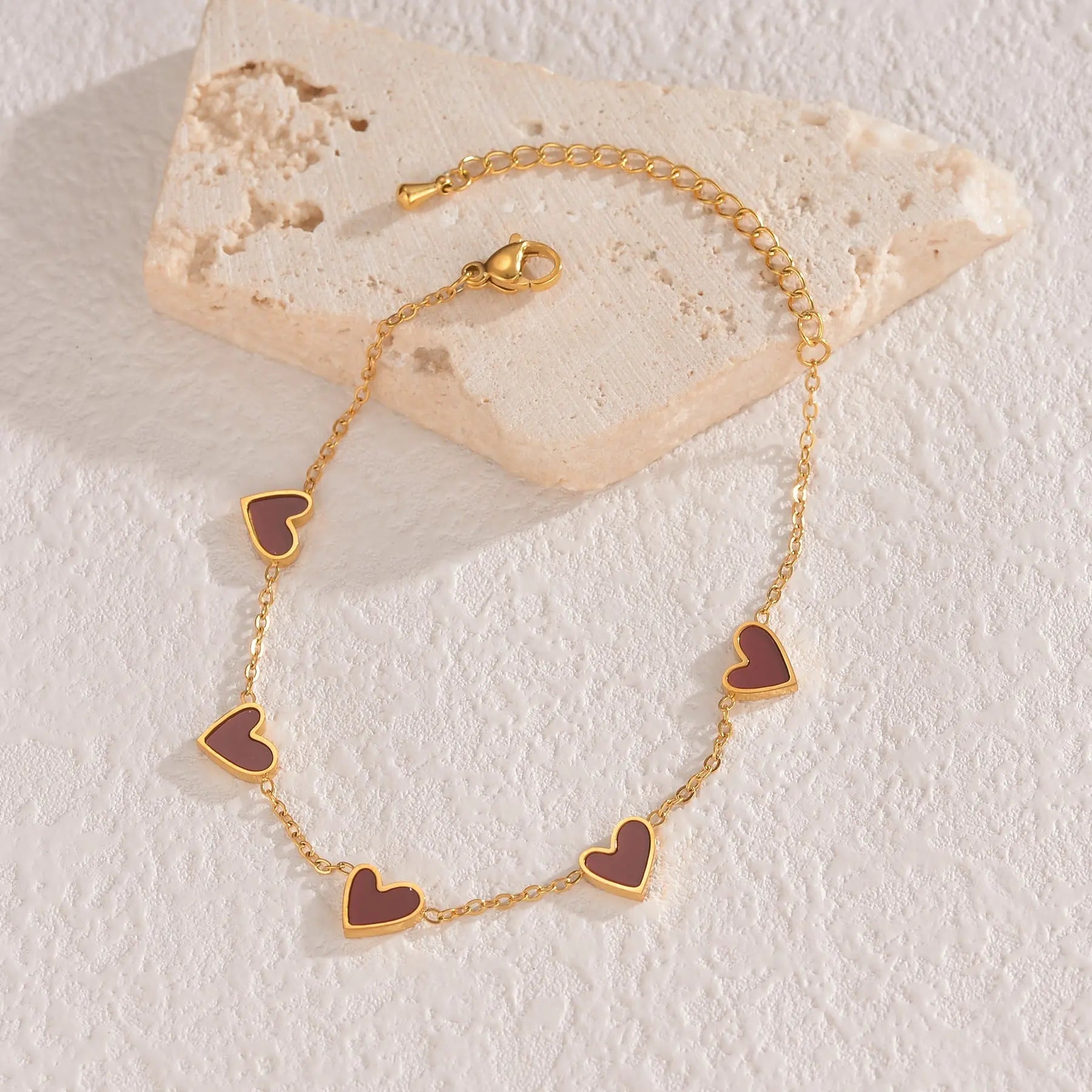 Gold-colored love heart bracelet with chain and heart charms on beige stone background