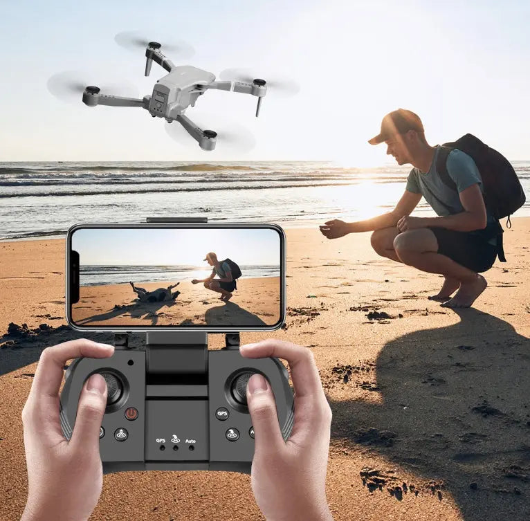 4K folding drone flying over beach controlled by remote with smartphone display showing aerial view of person and dog on sand at sunset