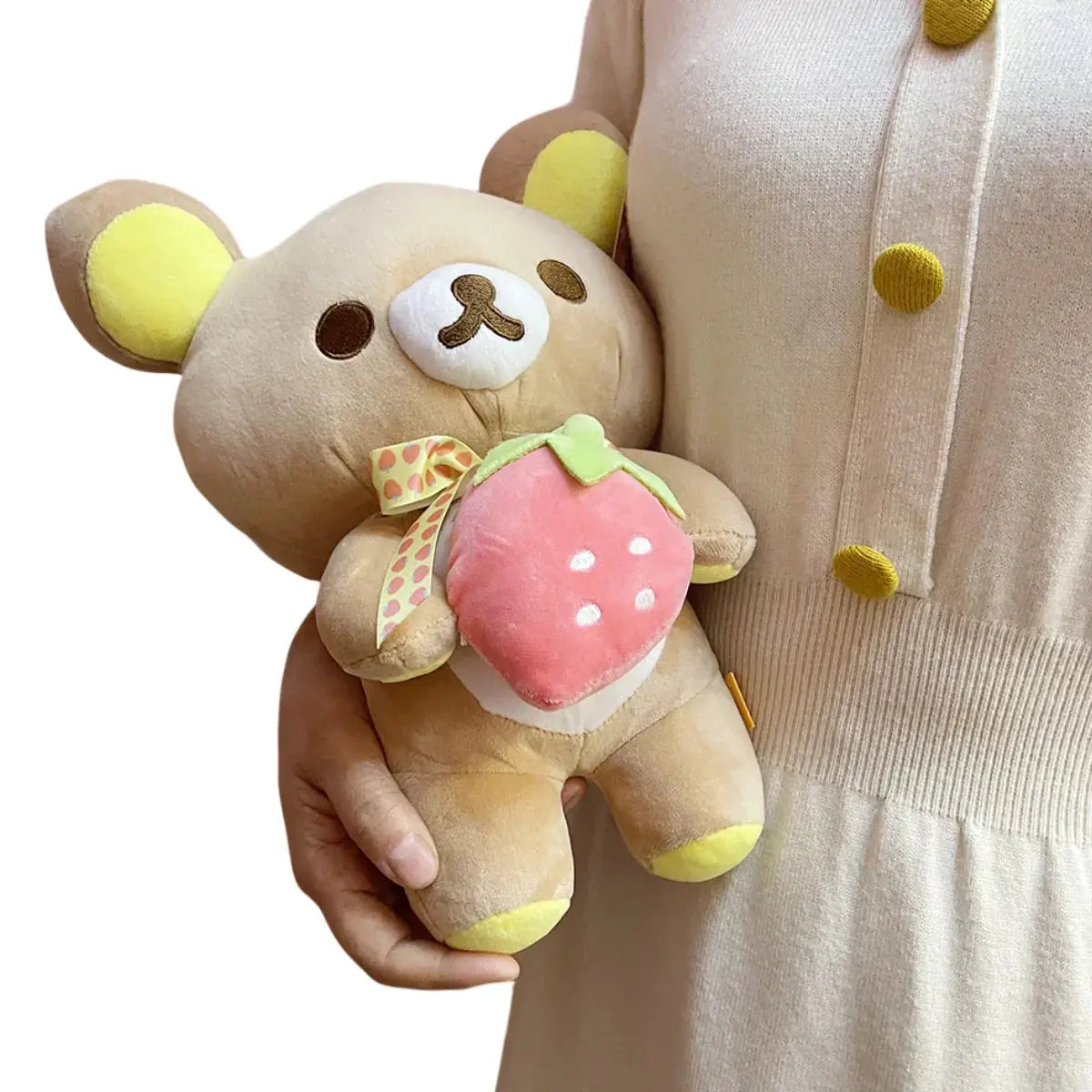 Plush toy bear holding a strawberry with a person wearing a polka dot shirt in the background