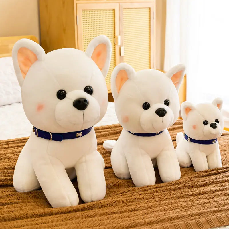 Three white plush puppy dolls in different sizes with blue collars on a brown bedspread in a cozy room